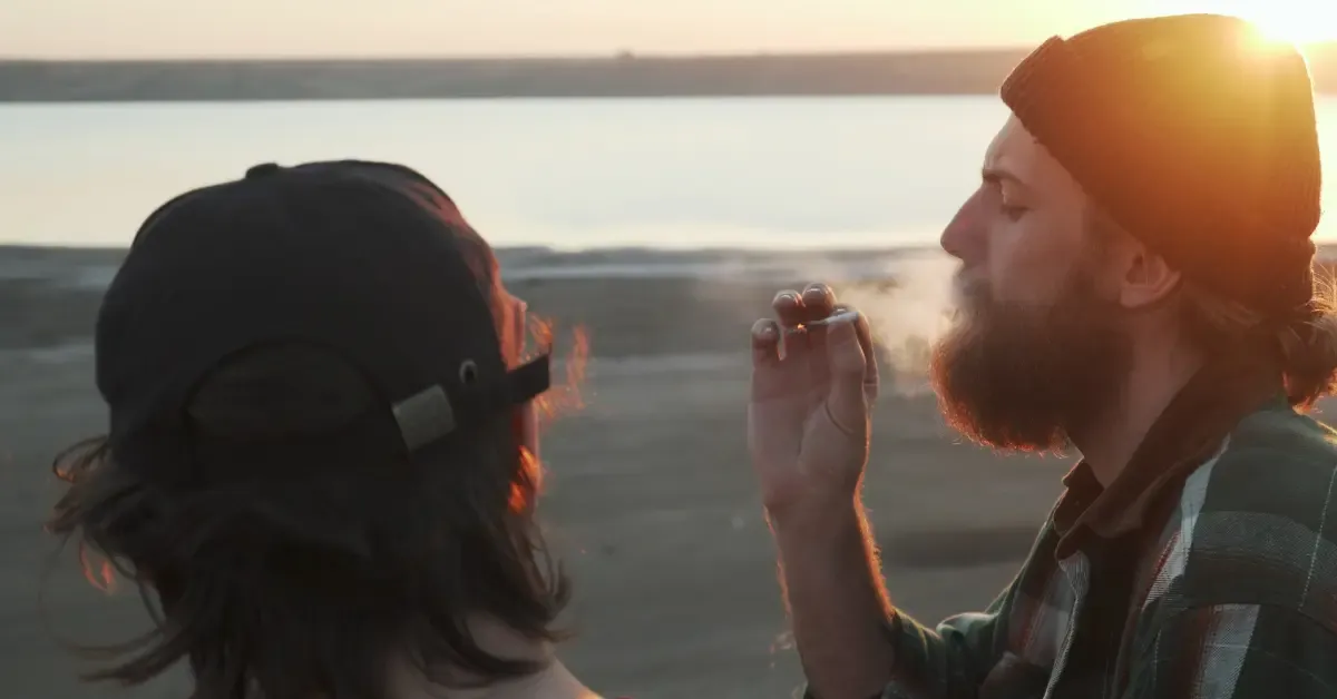 Two adult men smoking cannabis outdoors by the water at sunset