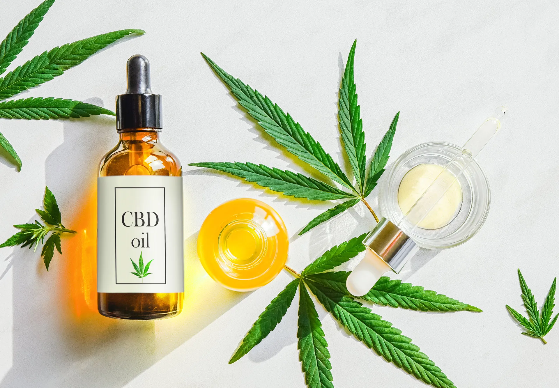 Amber dropper bottle labeled CBD oil beside a glass dropper, a dish of golden oil and fresh cannabis leaves on a white surface