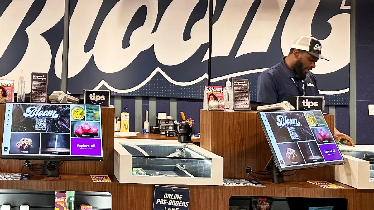 Budtender standing behind a Bloom dispensary counter with product display screens and a large Bloom wall logo