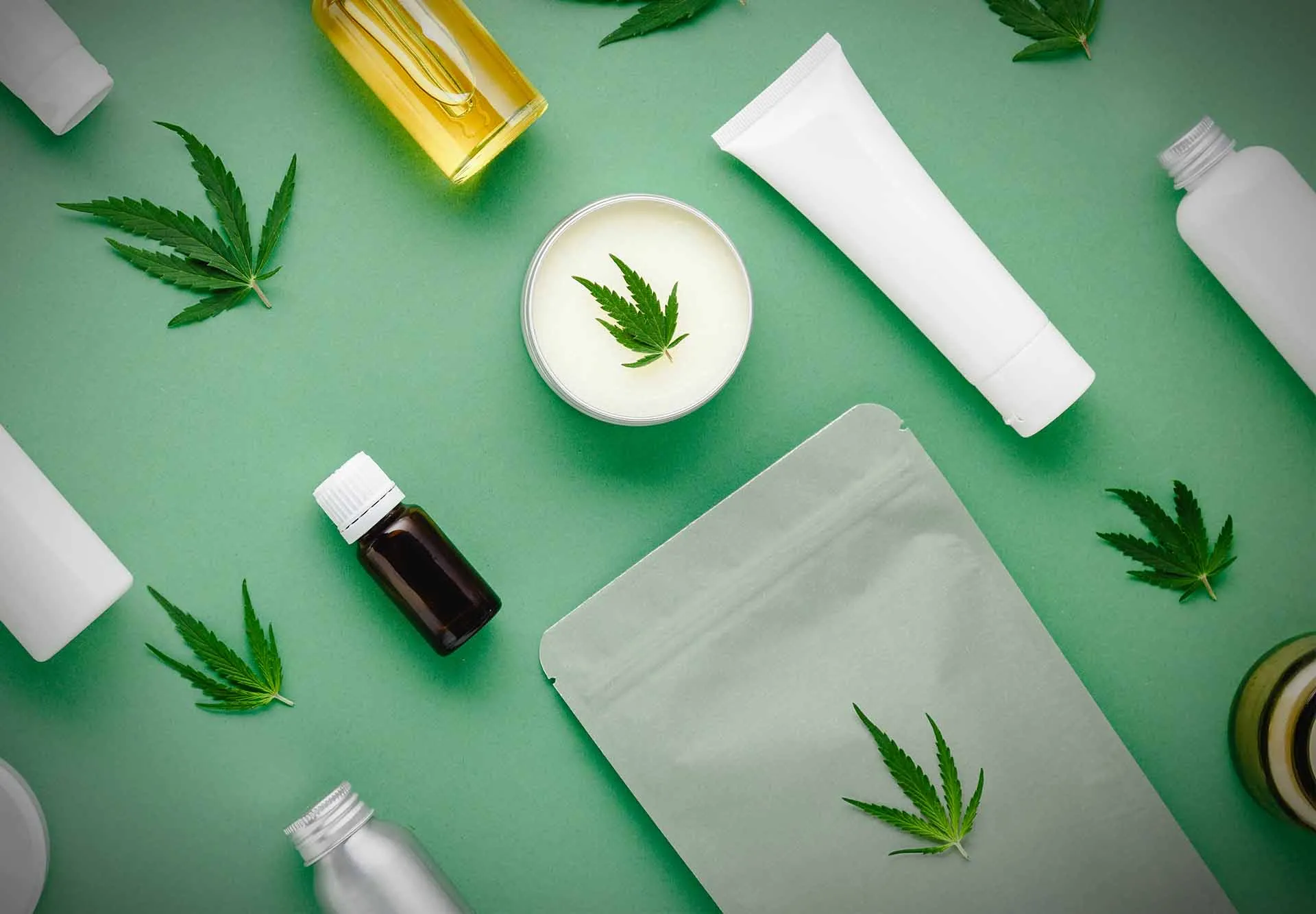 Unlabeled cannabis skincare products, including a cream jar, tubes, dropper bottles and an oil, laid flat with cannabis leaves on a green background