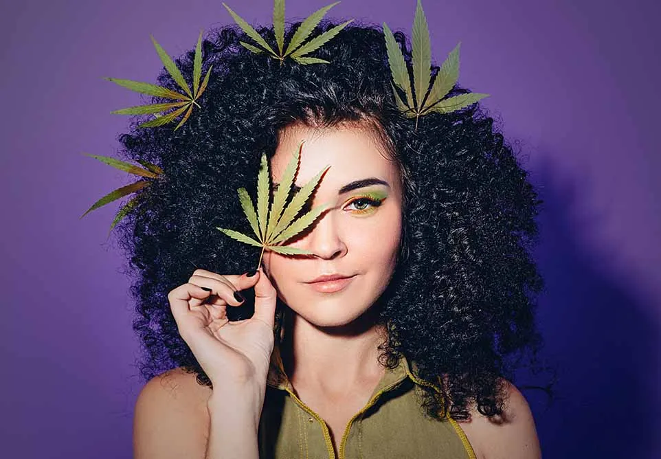 Woman with curly hair holding a cannabis leaf over one eye, with more leaves tucked in her hair, against a purple background