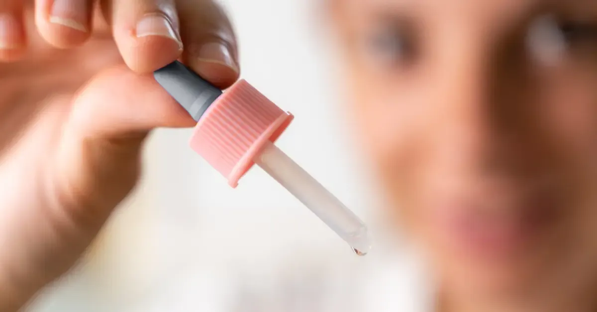 Hand holding a cannabis tincture dropper filled with oil, with a woman blurred in the background