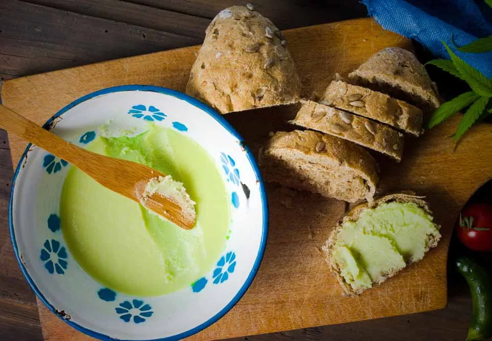 Bowl of green cannabis-infused butter with a wooden spreader beside sliced bread on a cutting board