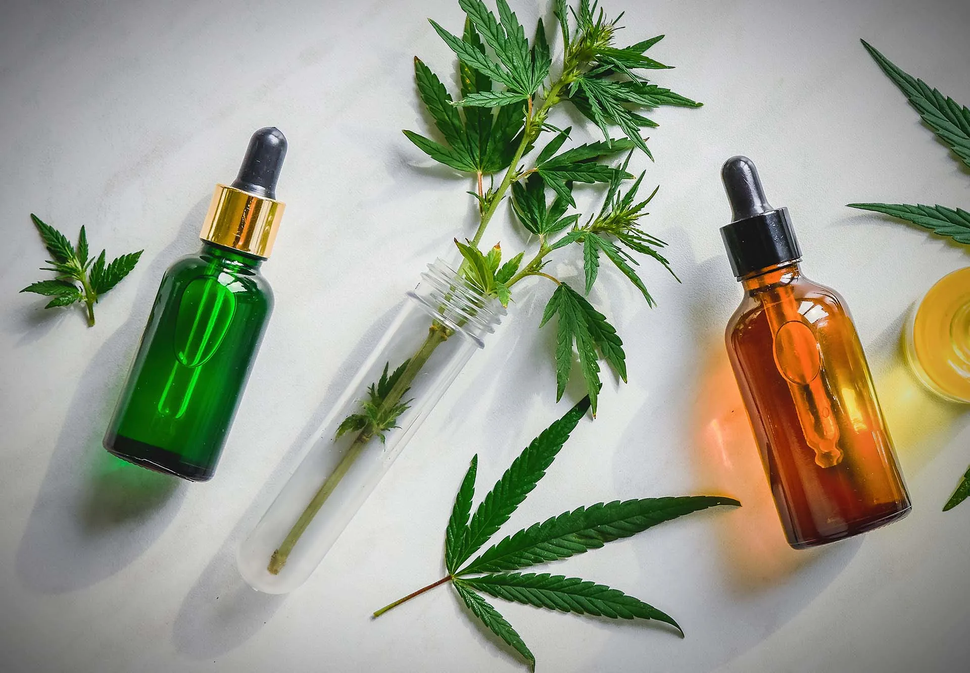Green and amber CBD tincture dropper bottles laid on a white surface with fresh cannabis leaves and a small glass vial