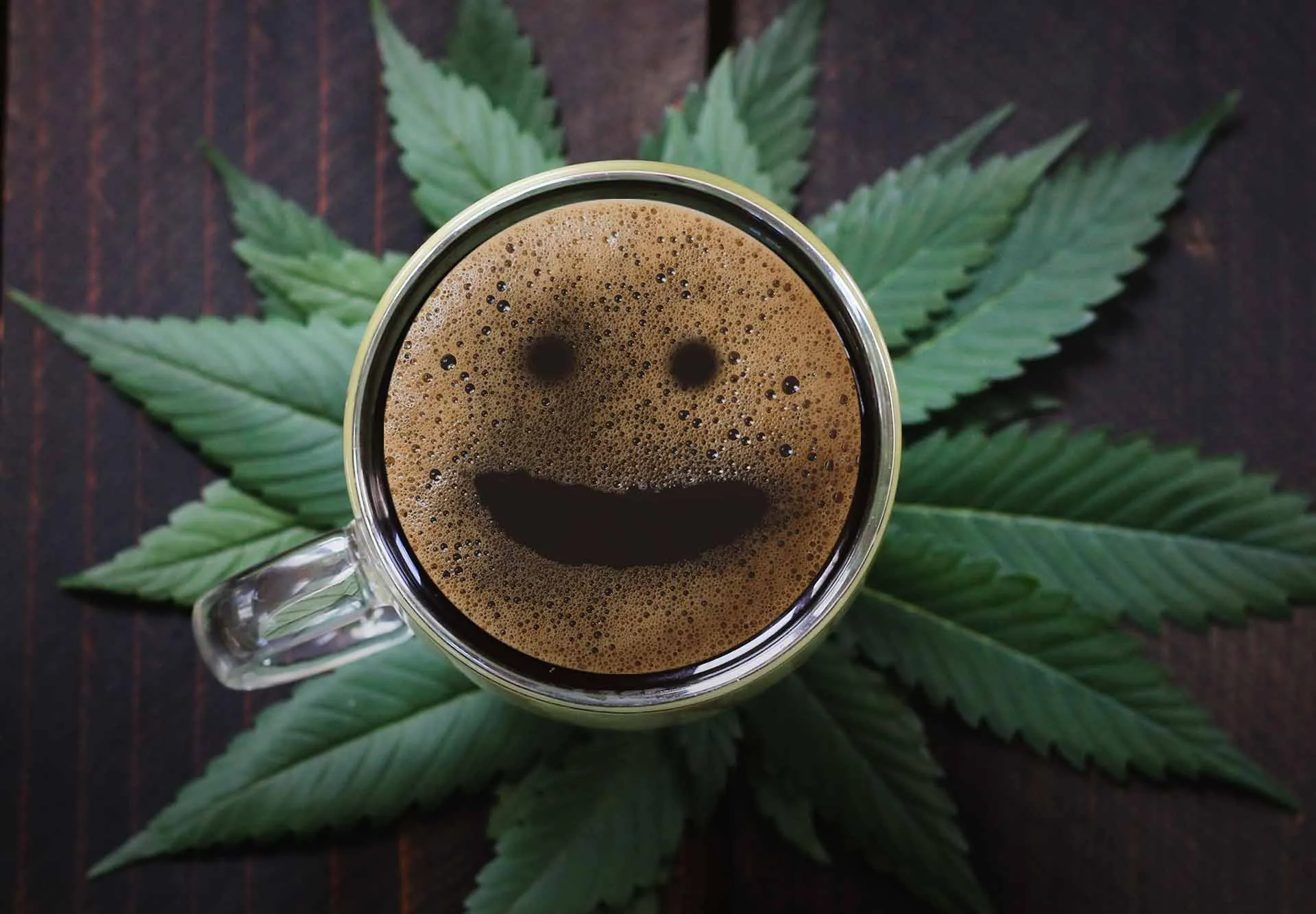 Glass cup of coffee with a smiley face in the foam sitting on cannabis leaves on a wooden table