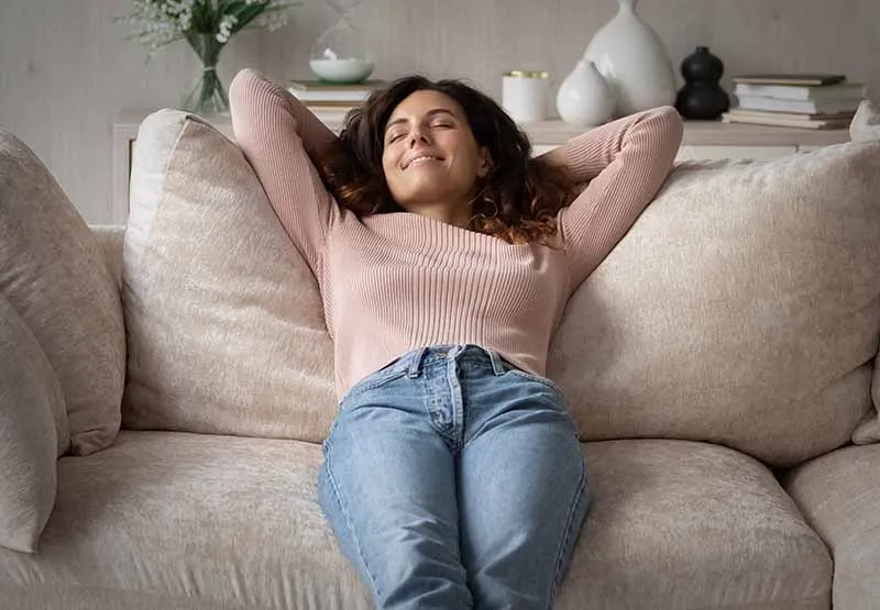 Woman in a pink sweater leaning back on a beige couch with her hands behind her head, eyes closed and relaxed