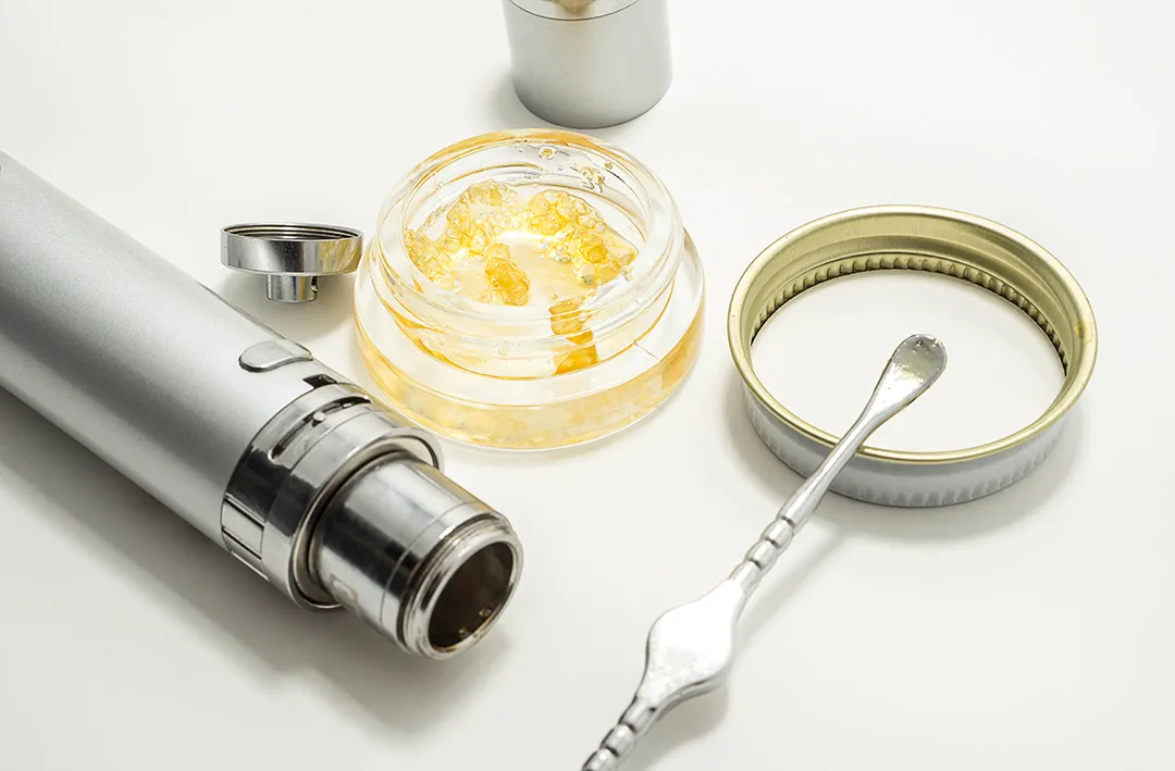 Silver vaporizer pen with its cap off beside an open glass jar of golden cannabis concentrate, a metal dab tool and a jar lid on a white surface