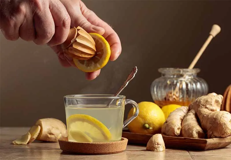 Hand squeezing a lemon over a glass of warm lemon water beside fresh ginger root and a jar of honey