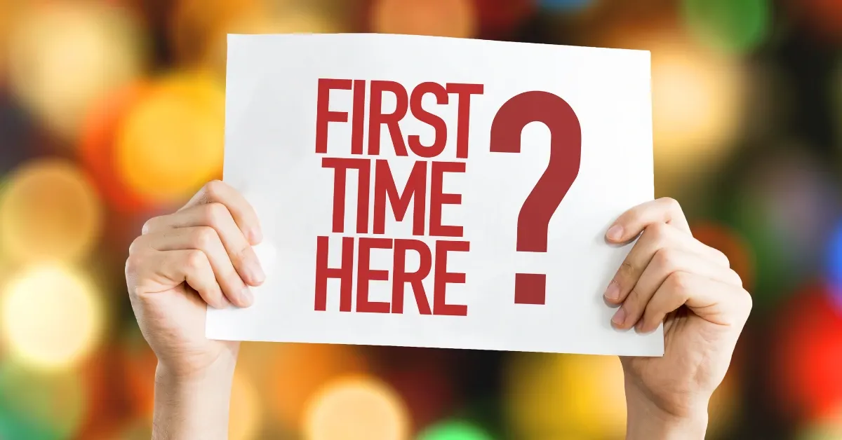 Hands holding up a white sign reading "First Time Here?" in red letters against a colorful blurred background