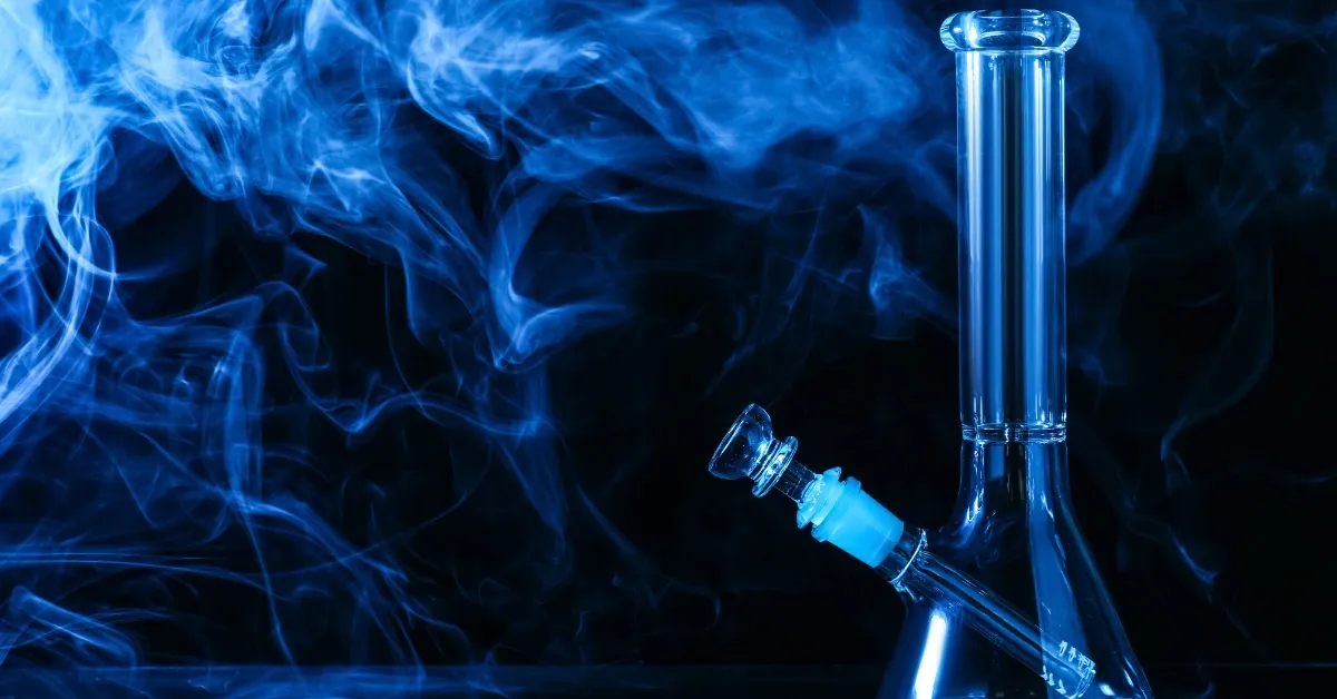 Clear glass beaker-base bong with a downstem and bowl, surrounded by swirling blue-lit smoke on a dark background