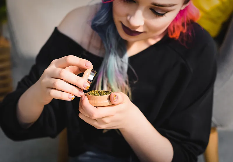 Woman with pastel-dyed hair holding a small wooden grinder filled with ground cannabis flower