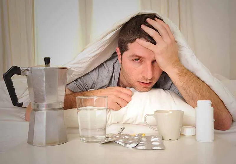 Hungover man in bed under a blanket holding his head beside a glass of water, pills, coffee pot and cup