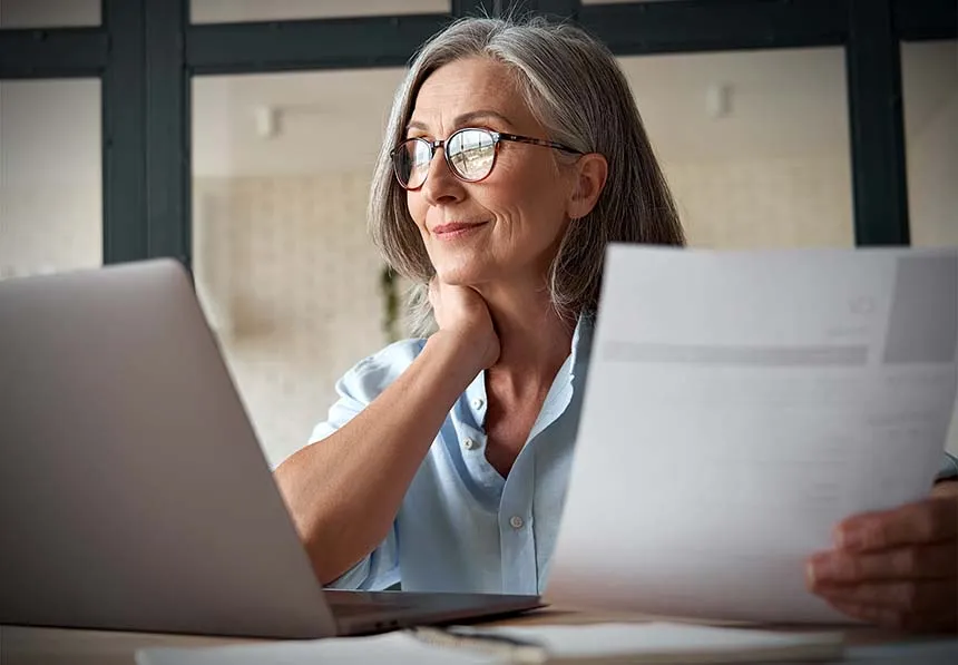 Older woman with glasses smiling at a laptop while holding paperwork at her desk