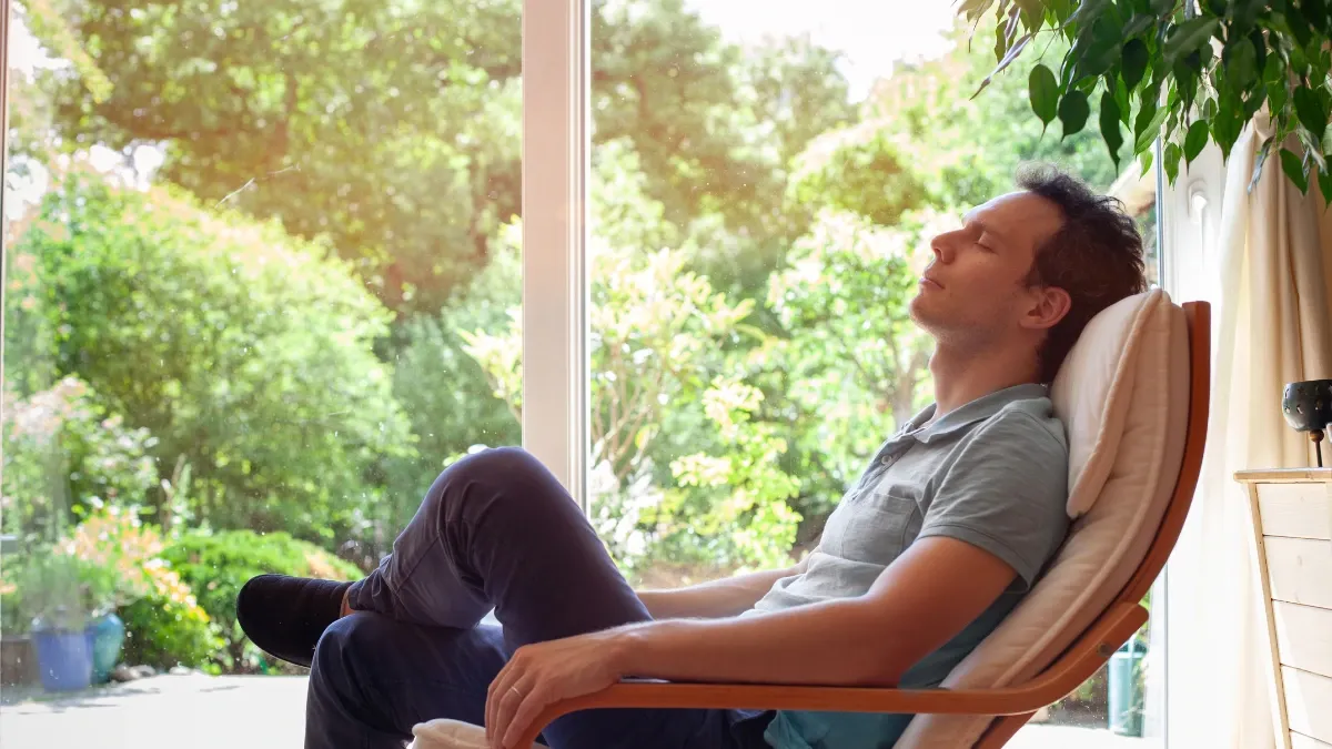 Man resting with his eyes closed in a lounge chair beside large windows overlooking a green garden