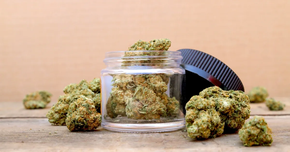 Open glass jar of cannabis flower buds with its black lid beside it, surrounded by loose buds on a wooden table