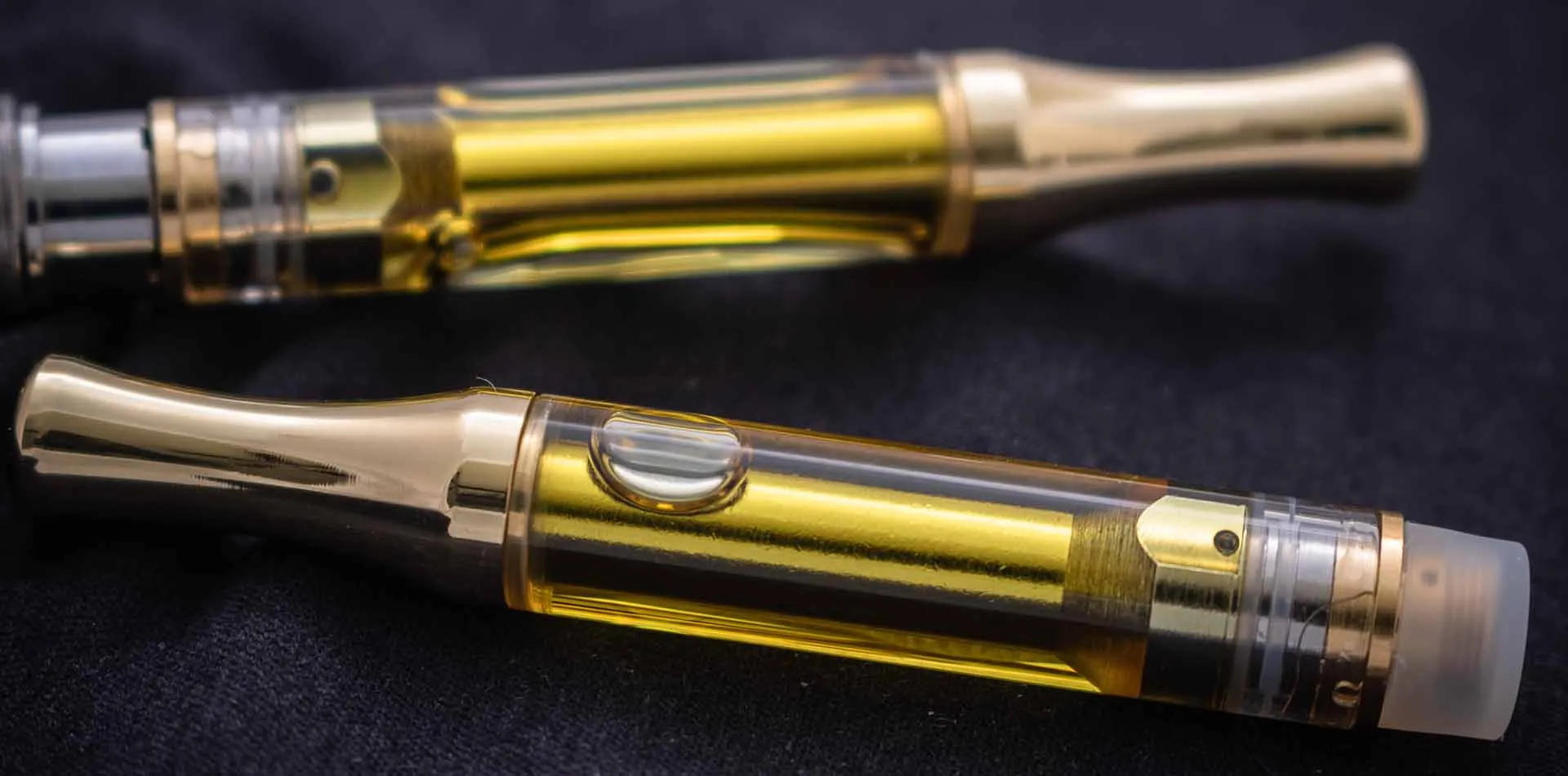 Two metal-tipped vape cartridges filled with golden cannabis oil on a dark background