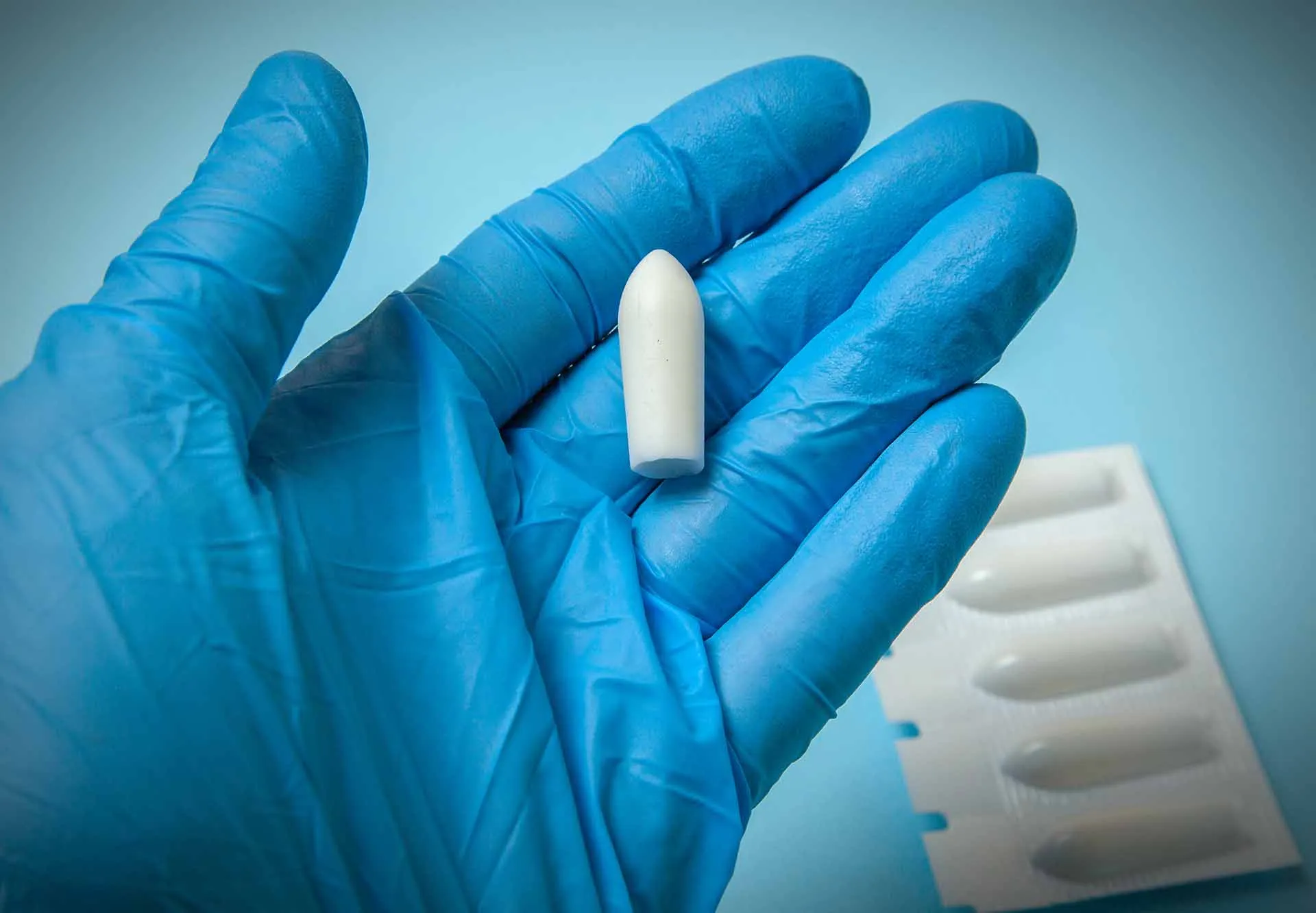 Gloved hand holding a white suppository in front of a blister pack of suppositories