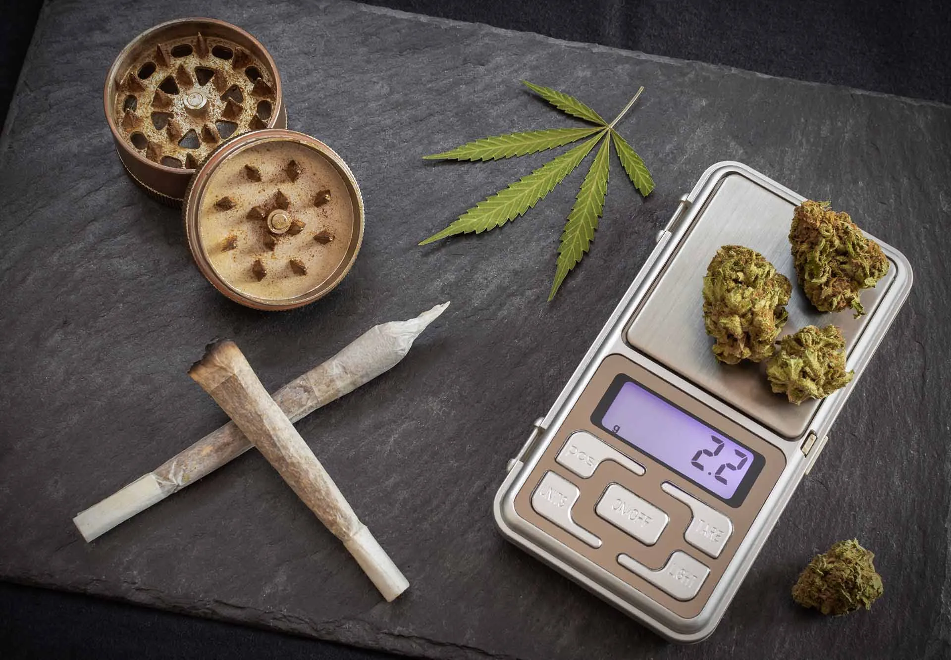 Cannabis buds on a digital pocket scale beside two pre-rolled joints, an open grinder and a cannabis leaf on a slate board