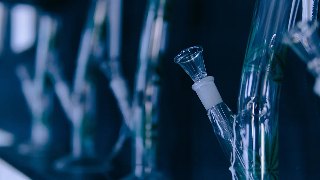Row of clear glass water pipes and bongs with glass bowl pieces on a display shelf in cool blue light