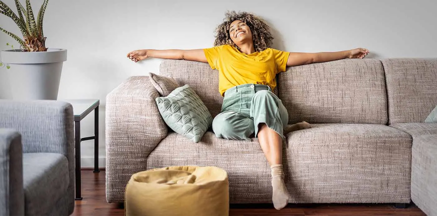 Smiling woman stretched out with arms spread wide on a gray sofa, sinking into a deeply relaxed couch lock feeling