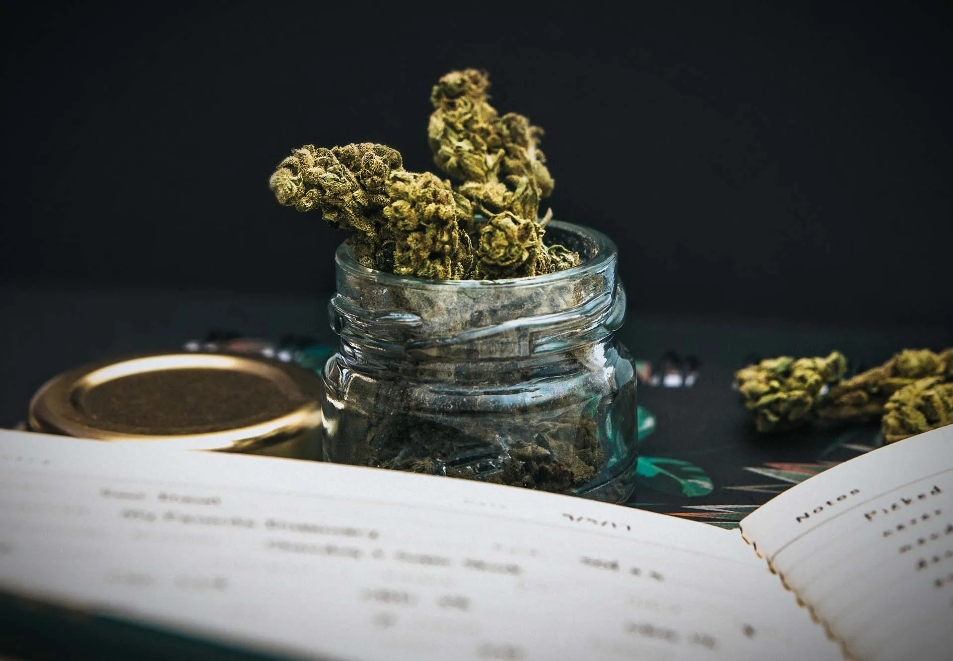 Glass jar of dried cannabis buds behind an open handwritten journal on a dark table