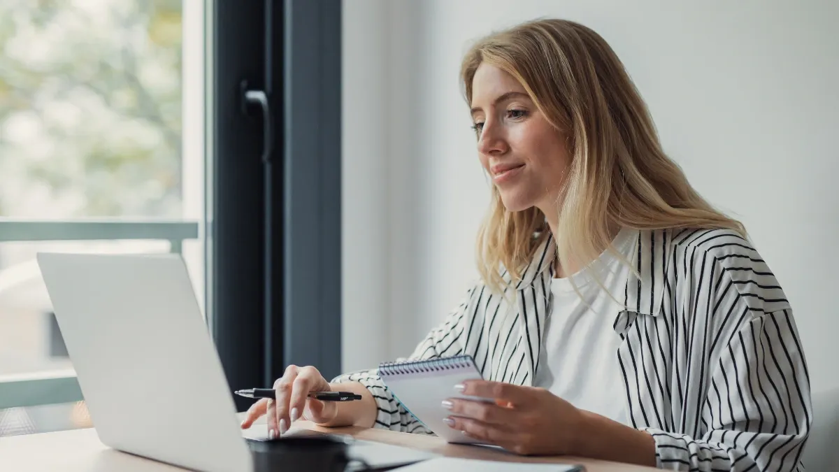 Woman working at a laptop by a sunny window, focused and holding a card